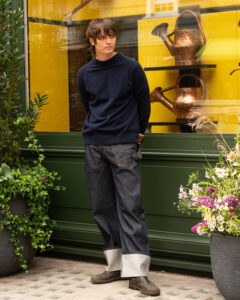 Man in a navy sweater and cuffed denim jeans stands outside a shop window with copper watering cans on display behind yellow panels