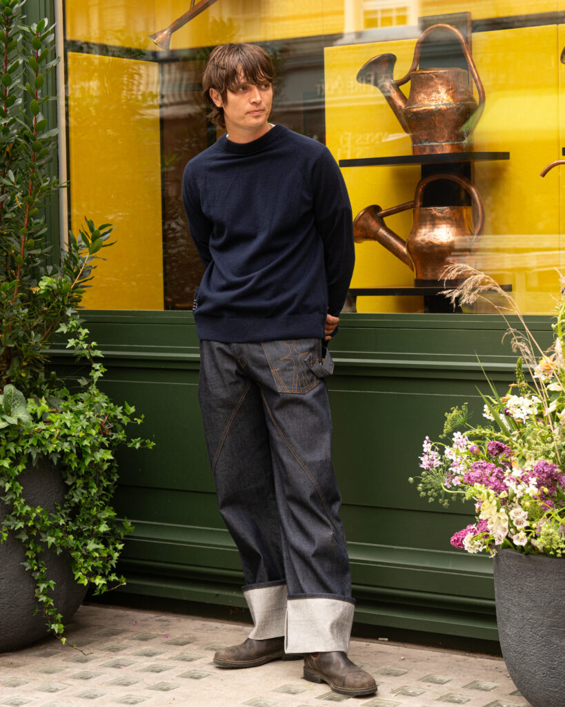 Man in a navy sweater and cuffed denim jeans stands outside a shop window with copper watering cans on display behind yellow panels