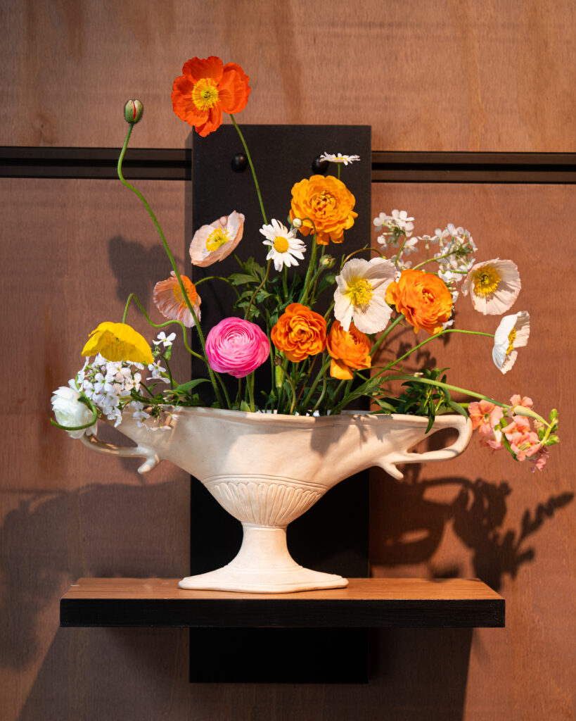 White pedestal vase holding a bright mixed bouquet of orange poppies, pink ranunculus, white daisies, and other small flowers against a wood wall backdrop.