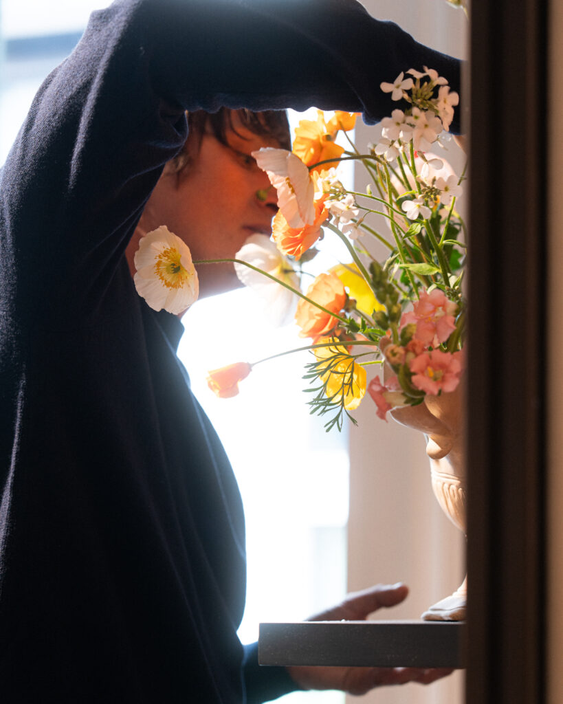 Person in a dark sweater hands a bouquet of orange and white flowers through a doorway.