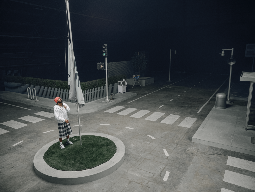 Amine in white hoodie and red turban holds a flagpole on a circular grassy island beside a crosswalk at night.