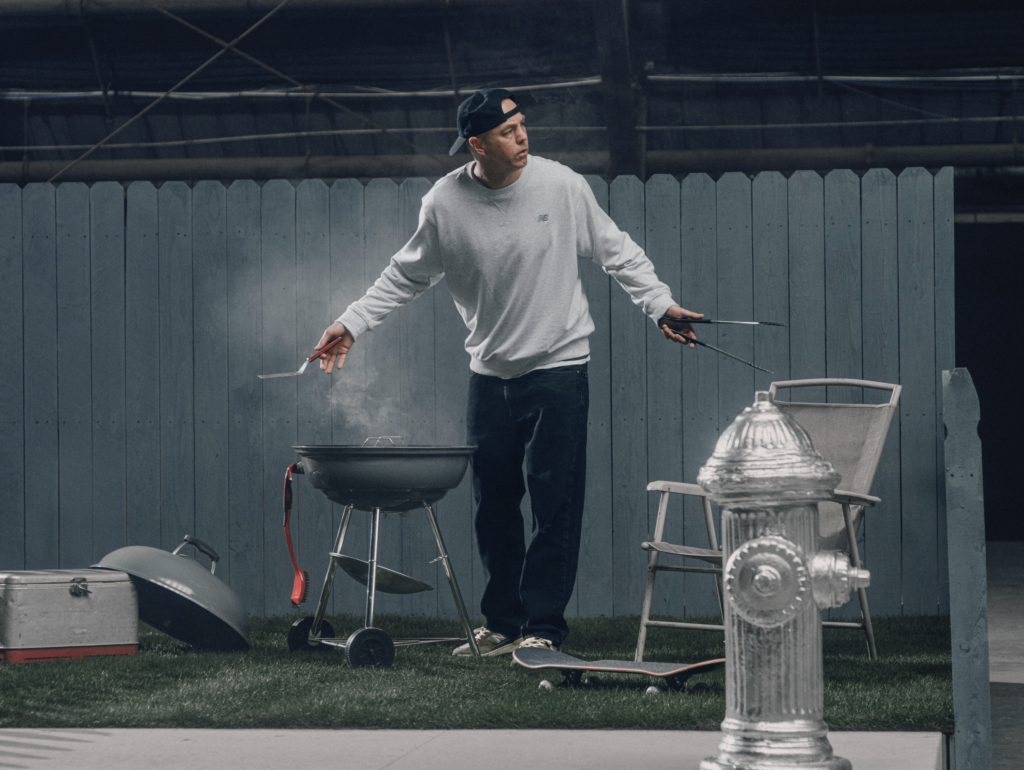 Andrew Reynolds wearing a gray sweatshirt and backward cap uses tongs at a backyard barbecue, with a grill and smoker nearby.