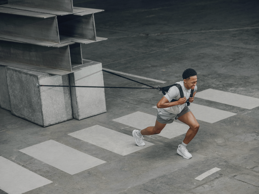 Quincy Wilson in athletic gear sprinting across a concrete plaza, tethered to a rope anchored to large concrete blocks on the left.