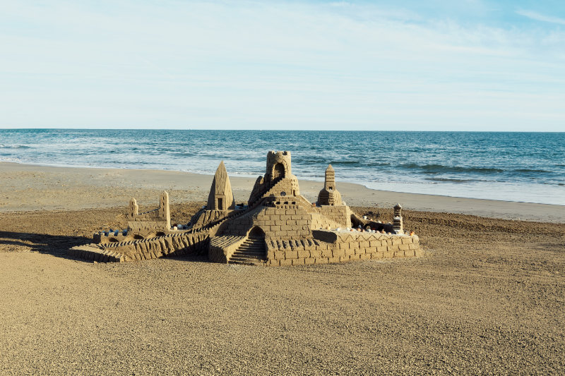 Sandcastle sculpture with towers on a sandy beach, ocean waves in the background at sunset-friendly light.