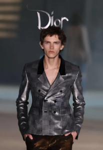 Male model walks the Dior runway wearing a metallic patchwork blazer with dark lapels and brown pants, hands in pockets.