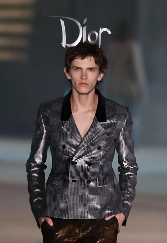 Male model walks the Dior runway wearing a metallic patchwork blazer with dark lapels and brown pants, hands in pockets.
