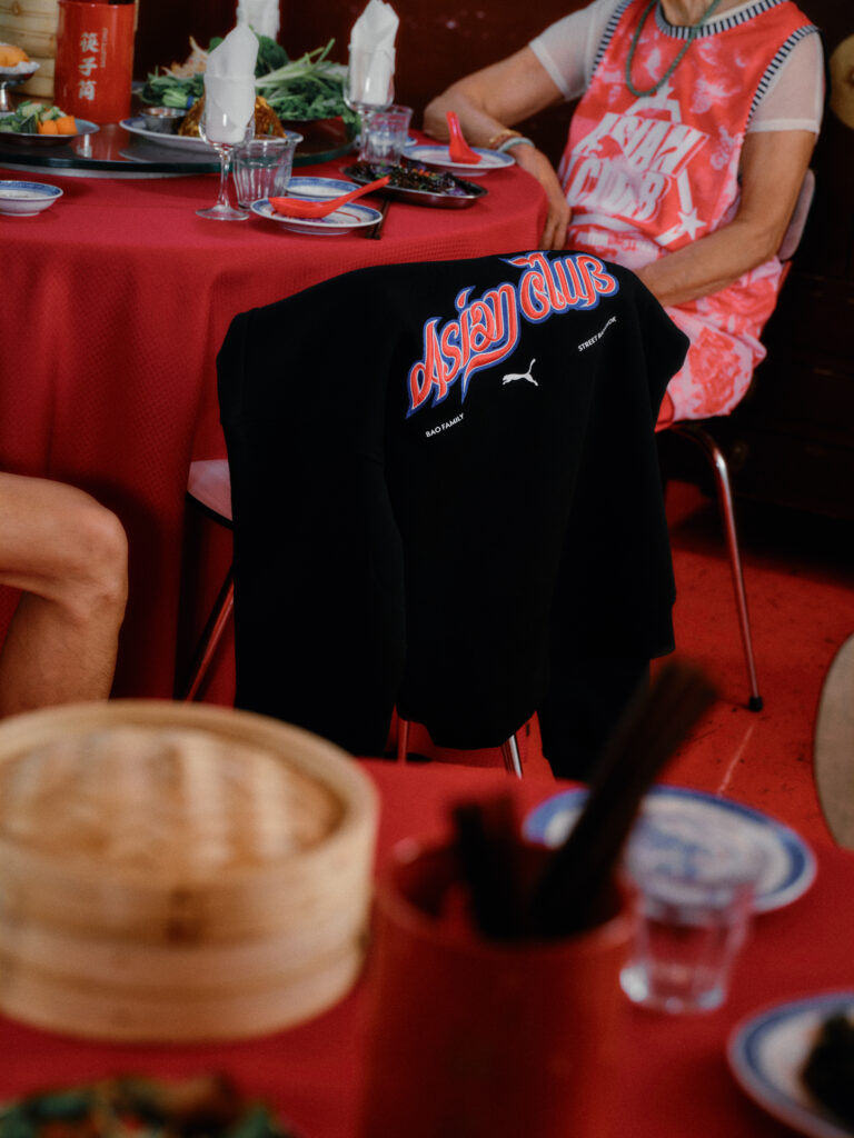 Person in a pink graphic shirt sits at a red table with dishes; a black jacket with blue-red 'Asian Club' logo is draped over the back of a chair.