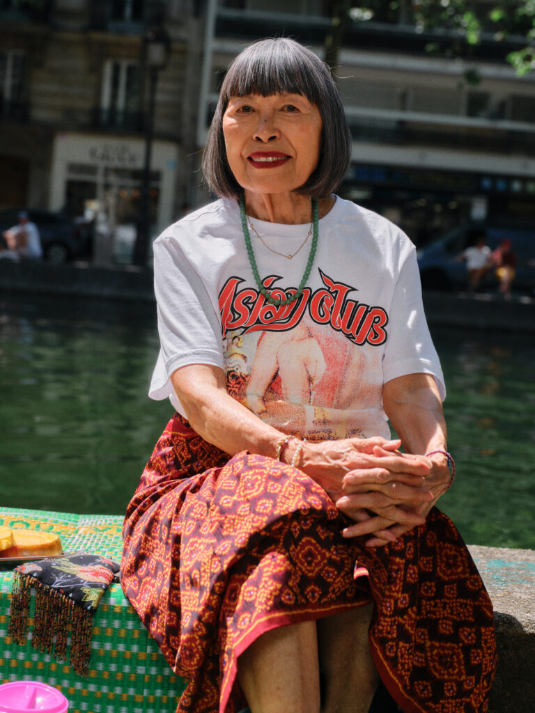 Smiling elderly woman with short gray hair sits by a waterway, wearing a white graphic T-shirt and green bead necklace.
