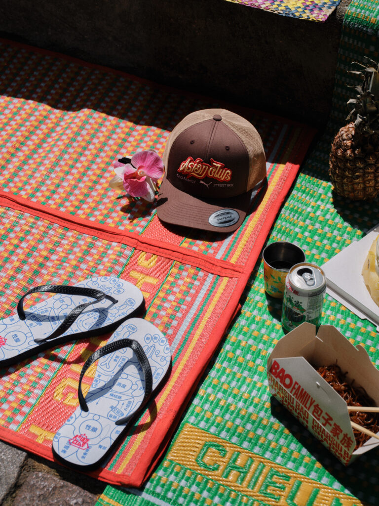 Brown trucker hat with red lettering placed on a colorful woven mat, orchid beside it, tropical market scene accents like pineapple and drinks nearby.