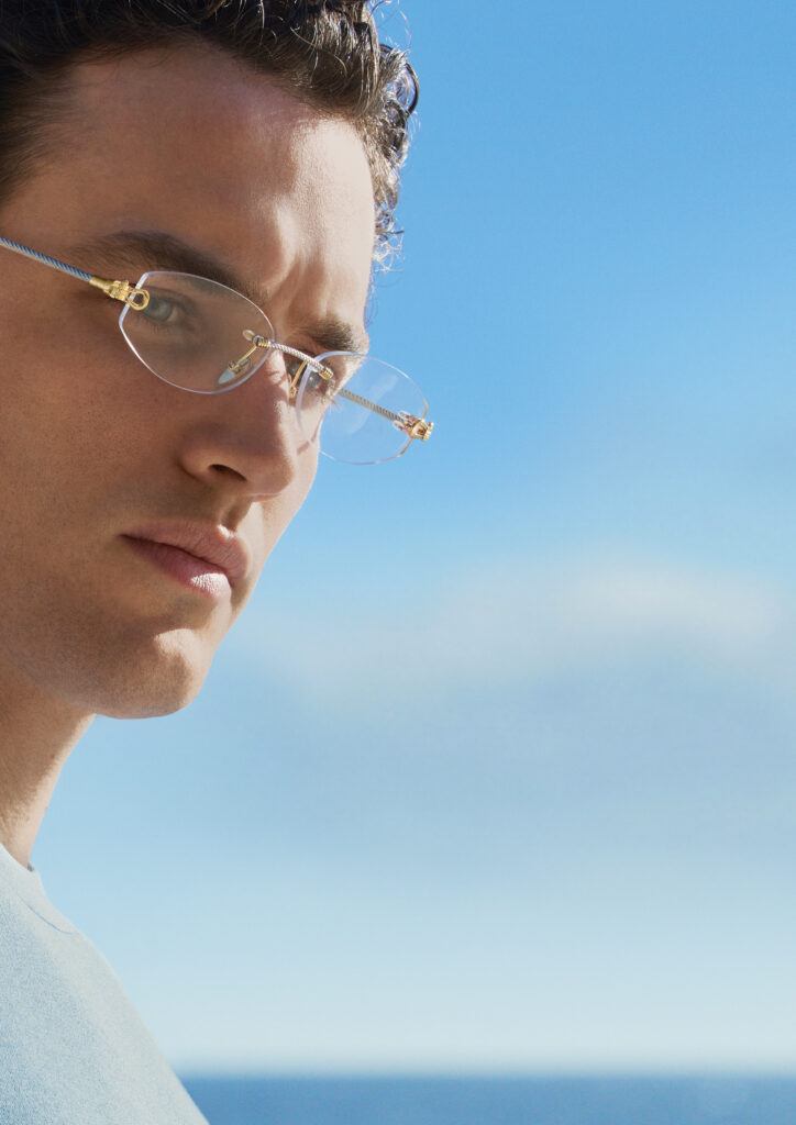 Close-up of a person wearing round clear FRED x THÉLIOS glasses, looking toward the horizon against a bright blue sky.