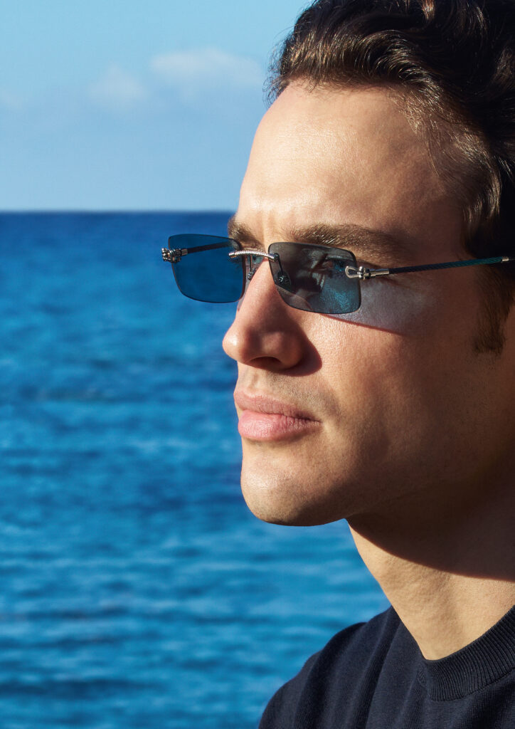 Close-up of a person wearing blue-tinted Fred x Thélios sunglasses with the ocean in the background.