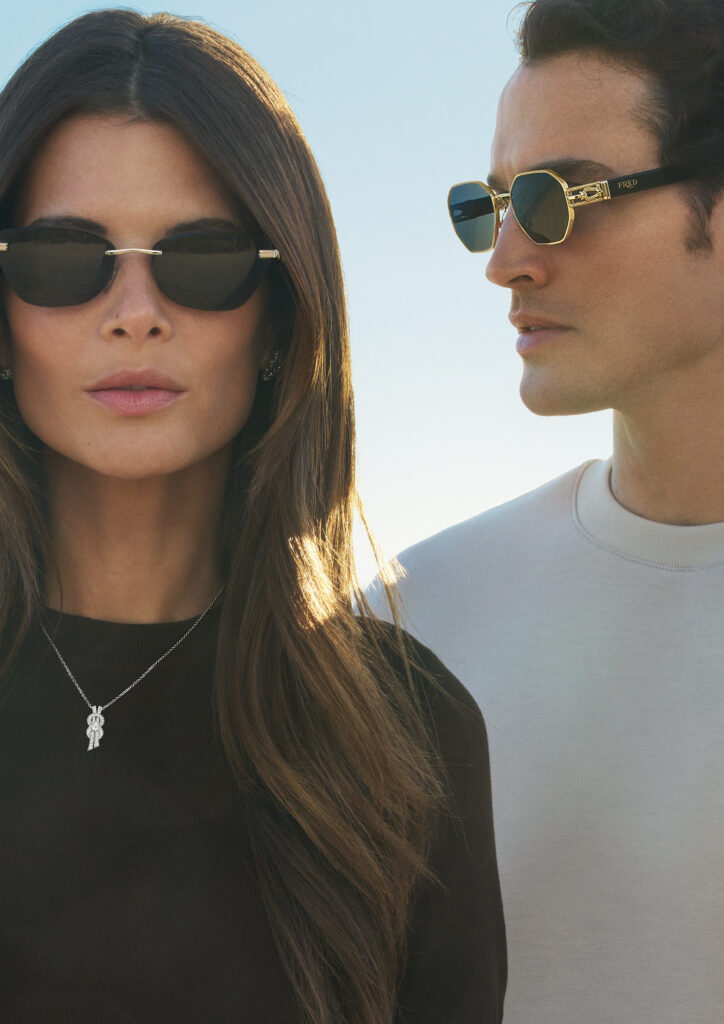 Two models wearing Fred x Thélios sunglasses: a woman in a black top with a pendant necklace on the left, and a man in a light shirt on the right, against a blue sky.