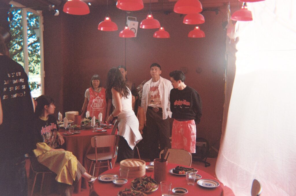 Group of people standing around a long red dining table with plates of food at a casual gathering indoors