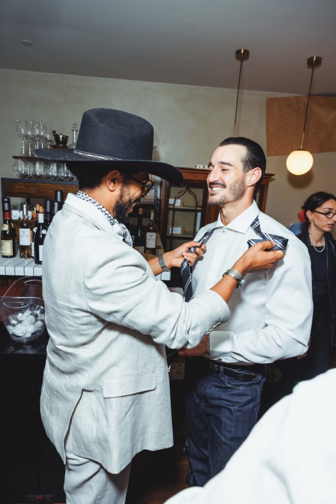 Two men at a bar: one in a light suit adjusts the other man's striped tie, smiling.