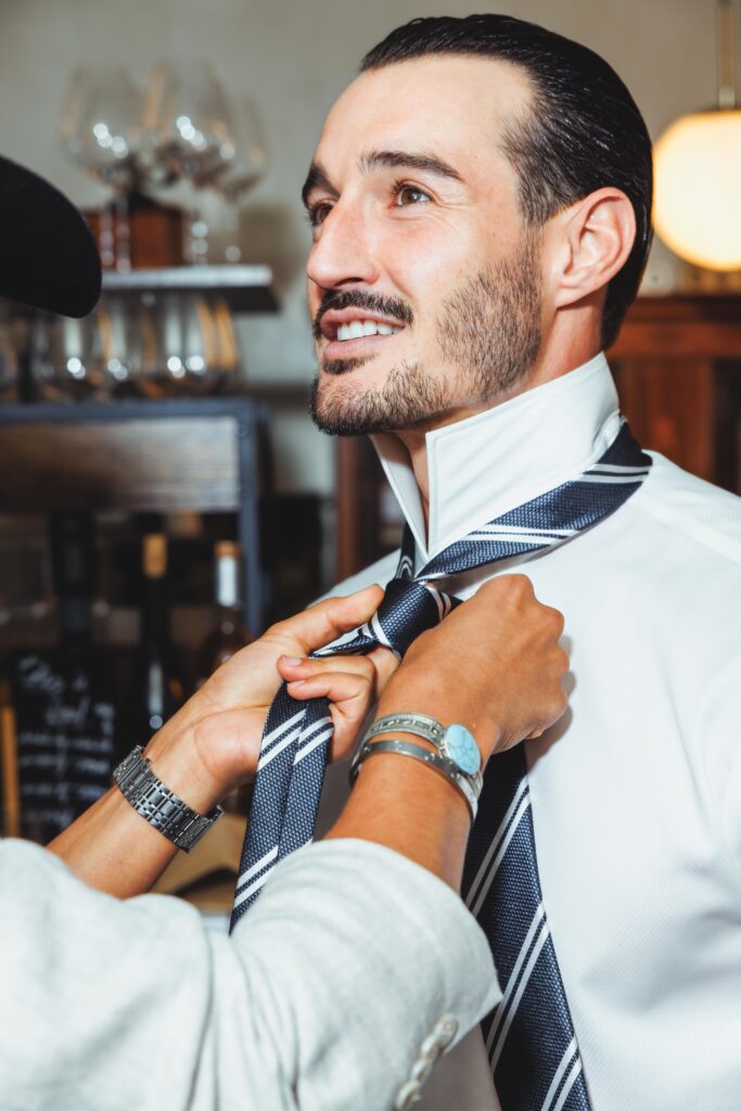 Man in a white shirt smiles as someone adjusts his striped navy tie in a cozy cafe setting.