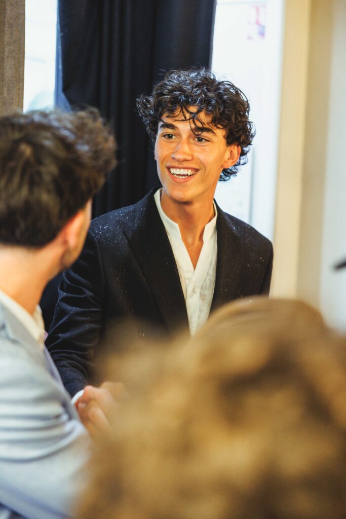 Young man with curly dark hair smiling while talking to friends in an indoor group setting.