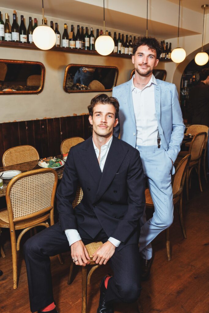 Two men in formal suits pose in a restaurant with bottles on a shelf and round hanging lights in the background.