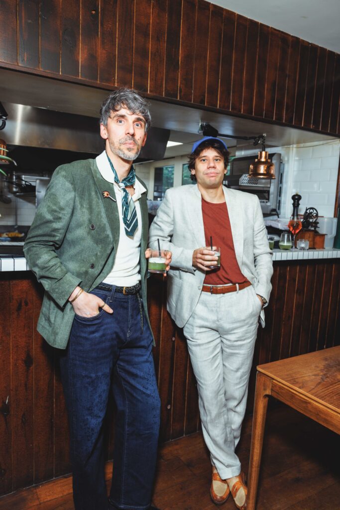 Two casually dressed men pose at a bar, each holding a drink; retro wooden paneling and copper lamp in the background.