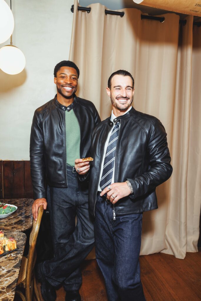 Two men in leather jackets smiling at a party, standing by a table of snacks in a home setting.
