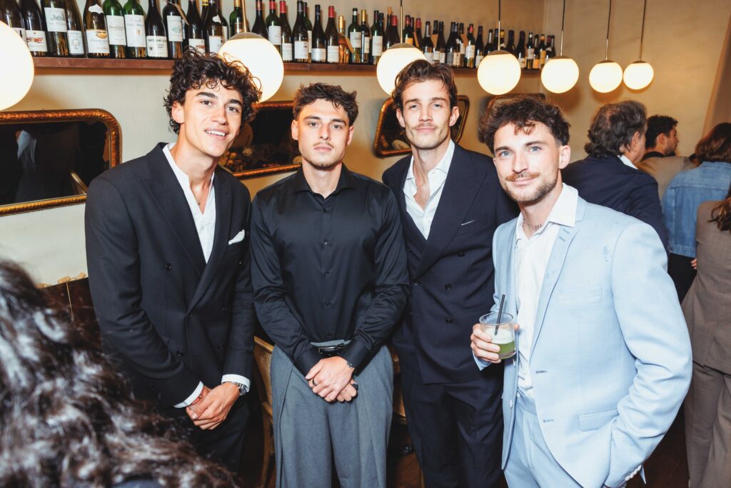 Four men in suits posing at a bar with wine bottles lining the shelf behind them.