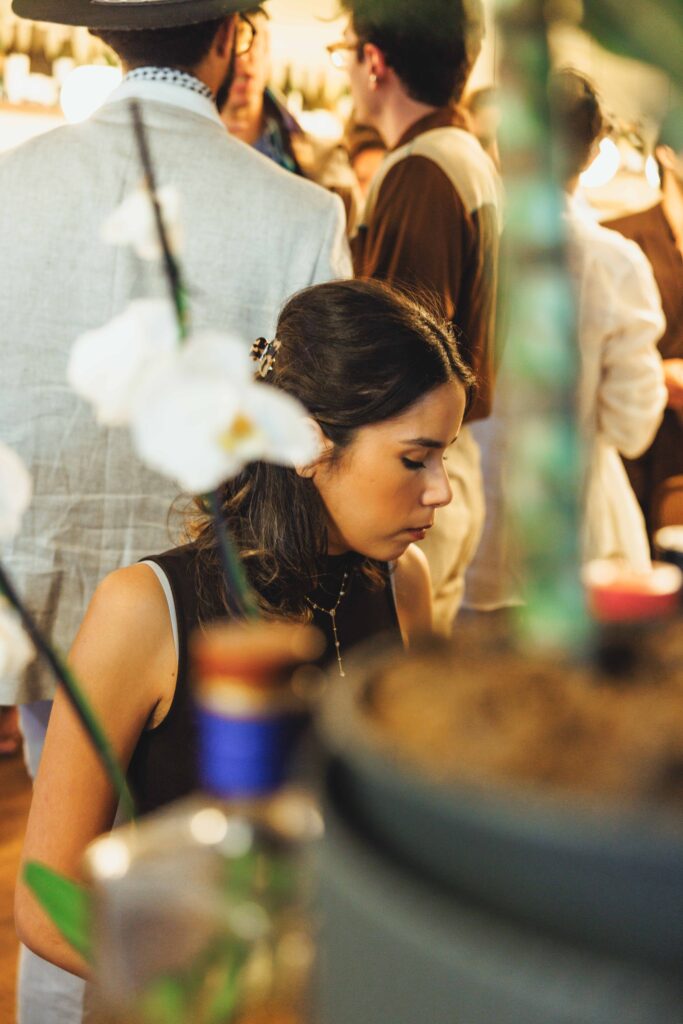 Woman with dark hair in a black top looking down, surrounded by people at a warm, blurred social gathering.