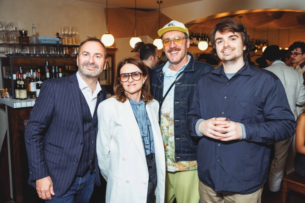 Four adults posing together at a bar, smiling for the camera with bottles and glassware in the background on shelves behind them