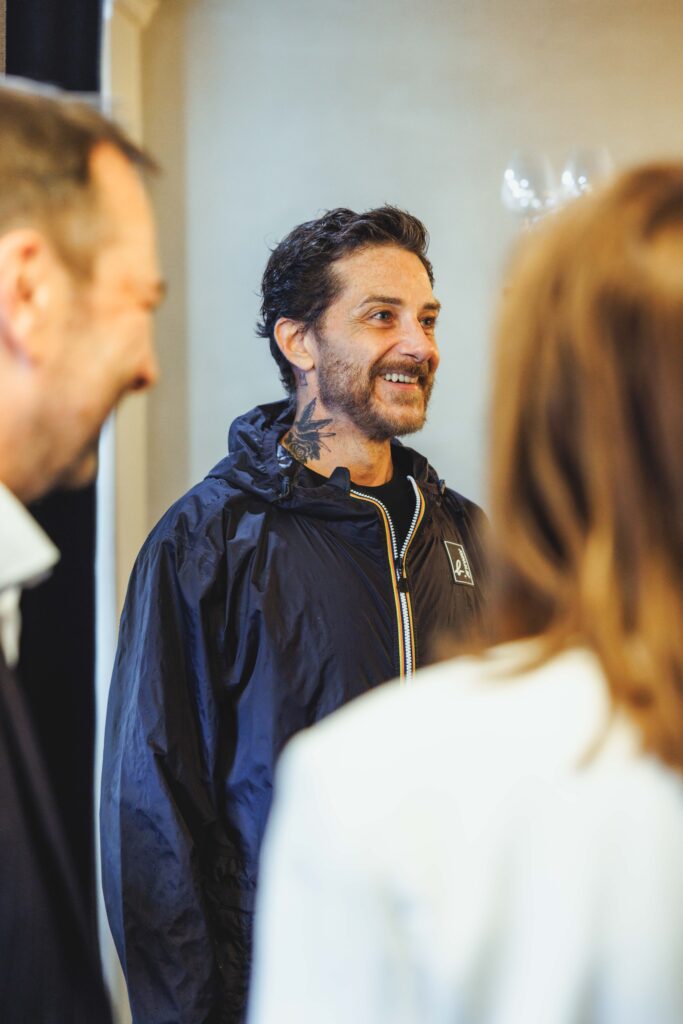 Smiling Sandro ACCARIÈS owner and editor in chief of Salut les Garçons Magazine with a neck tattoo and dark hair wearing a navy windbreaker, talking in a group.