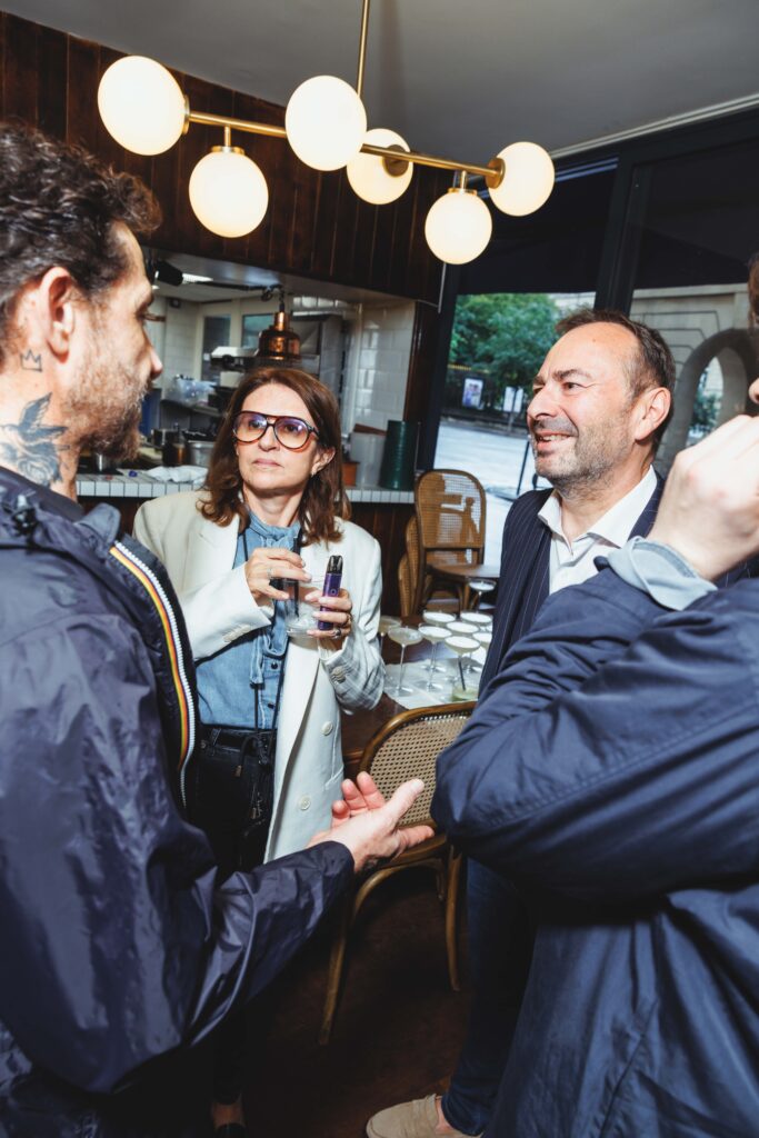 Three adults chat in a cafe near a bar, a woman with glasses holds a drink while a man in a suit smiles nearby, warm globe lights overhead.