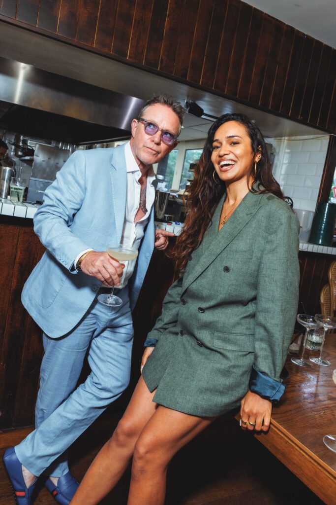 Two friends posing at a bar, smiling; man in a light blue suit holding a cocktail, woman in a green blazer dress beside him.
