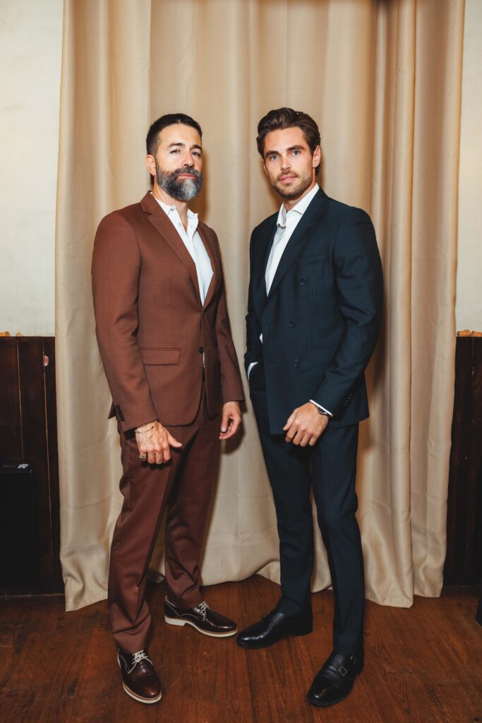 Two men in suits pose indoors in front of beige curtains; one in brown suit, the other in navy suit.