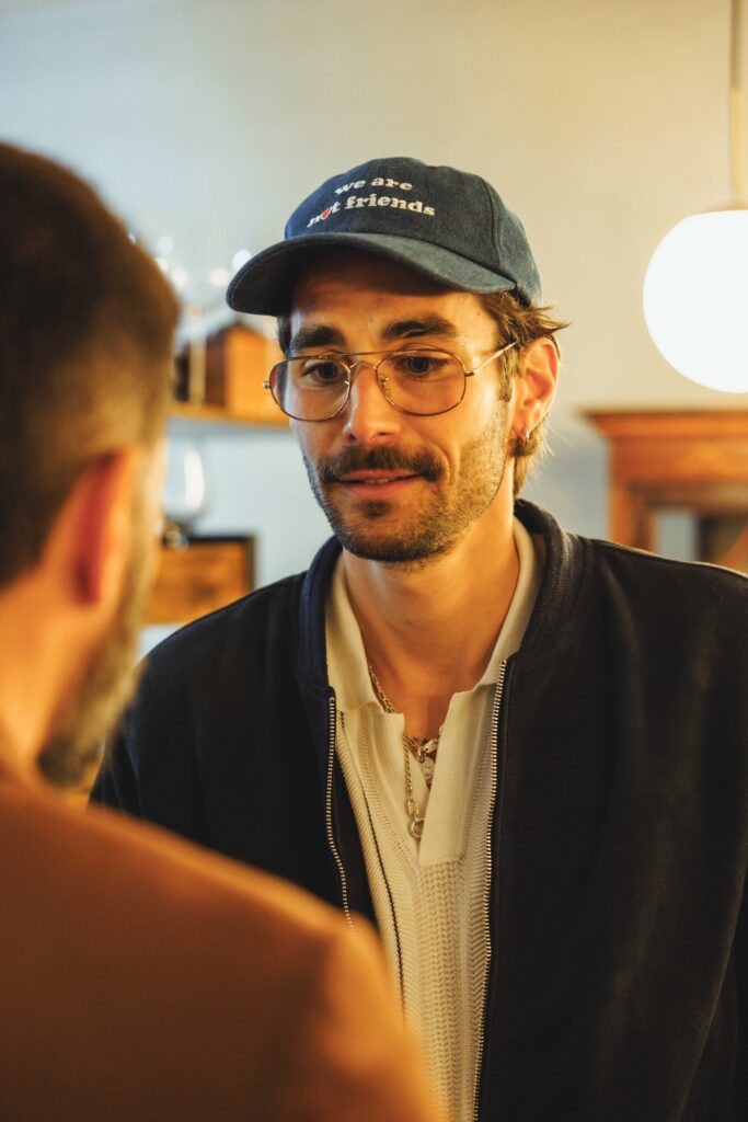 Man with glasses wearing a blue cap that says 'we are not friends' talks to someone indoors, close-up portrait style.
