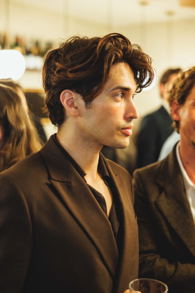 Side profile of a young man with dark hair wearing a brown blazer at a formal gathering, holding a glass in the foreground.