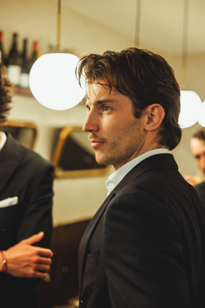Man in a dark suit in profile view at a social event, blurred bar/background in soft lighting.