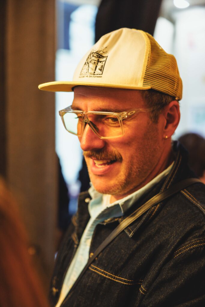 Man in a beige mesh-back cap and clear-framed glasses, smiling as he looks to the side indoors.