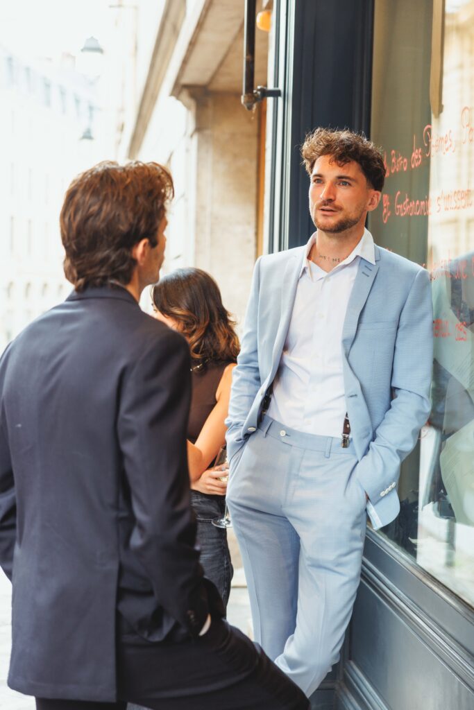 Two men in a street conversation outside a storefront; one man in a light-blue suit leans against the window as others chat nearby.