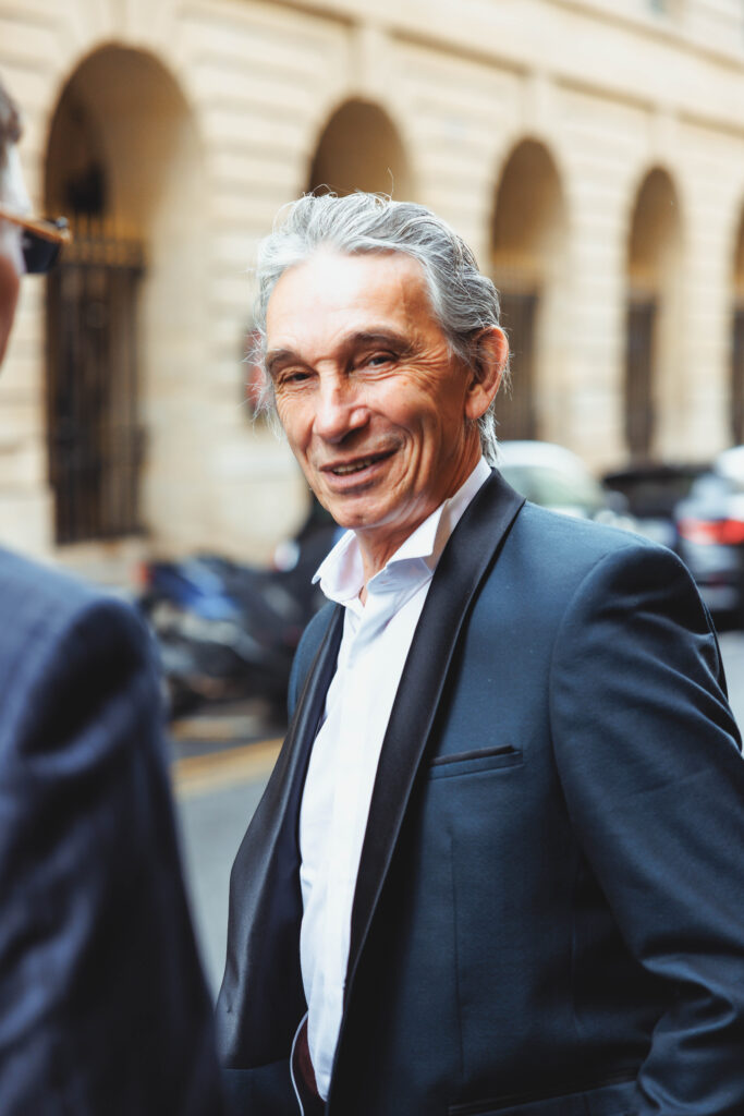 Smiling older man in a dark suit and white shirt, standing outdoors with arched stone buildings in the background
