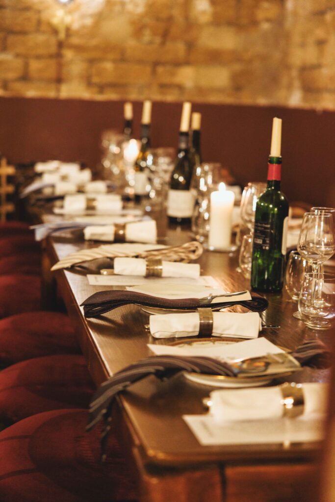 Long dining table in a cozy restaurant set for a meal with folded napkins, silverware, wine bottles, candles, and glassware in view near a brick wall.