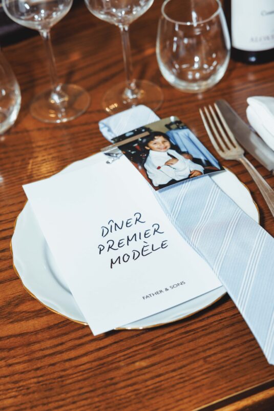 Table setting with a white menu card reading 'DINER PREMIER MODELE' on a wooden table, flanked by wine glasses and cutlery with a blue napkin nearby