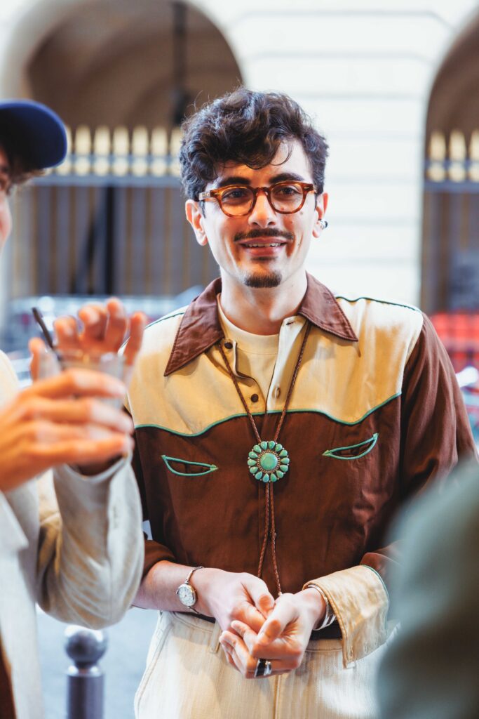 Man with brown round glasses and a dark mustache wearing a brown-and-cream western-style shirt and turquoise pendant, smiling in a social setting.