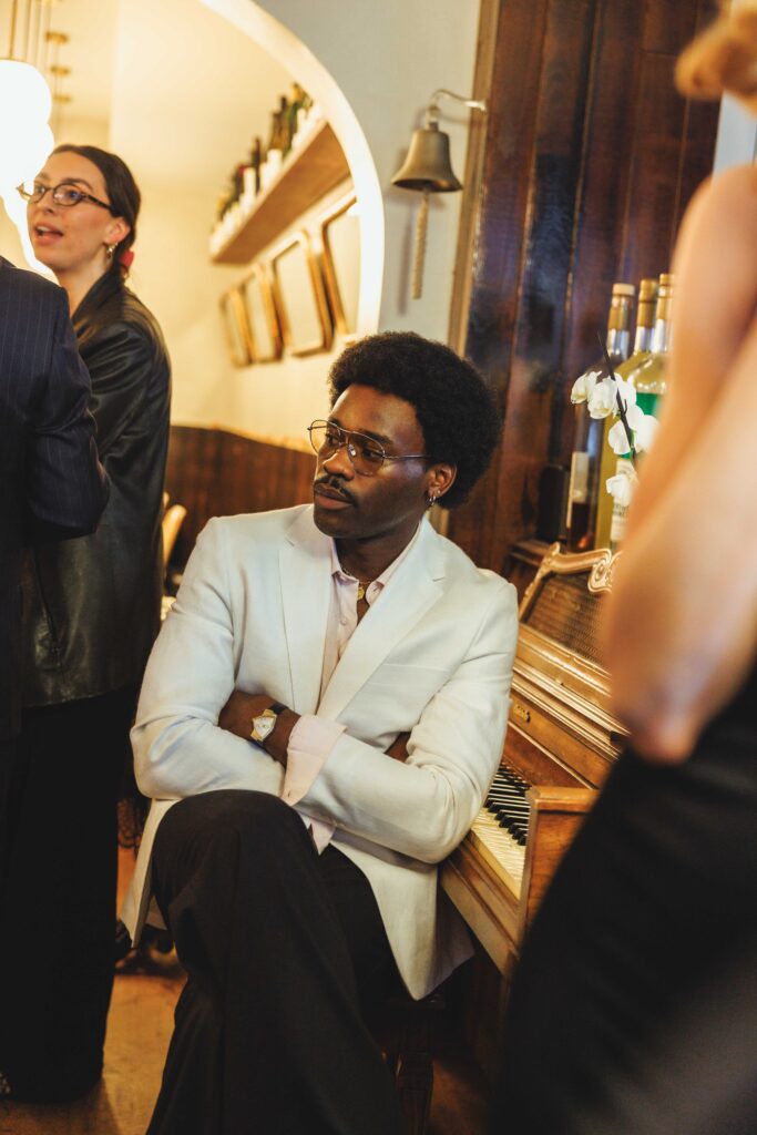 Man in a light blazer sitting by a piano in a warmly lit bar, with other people standing nearby.