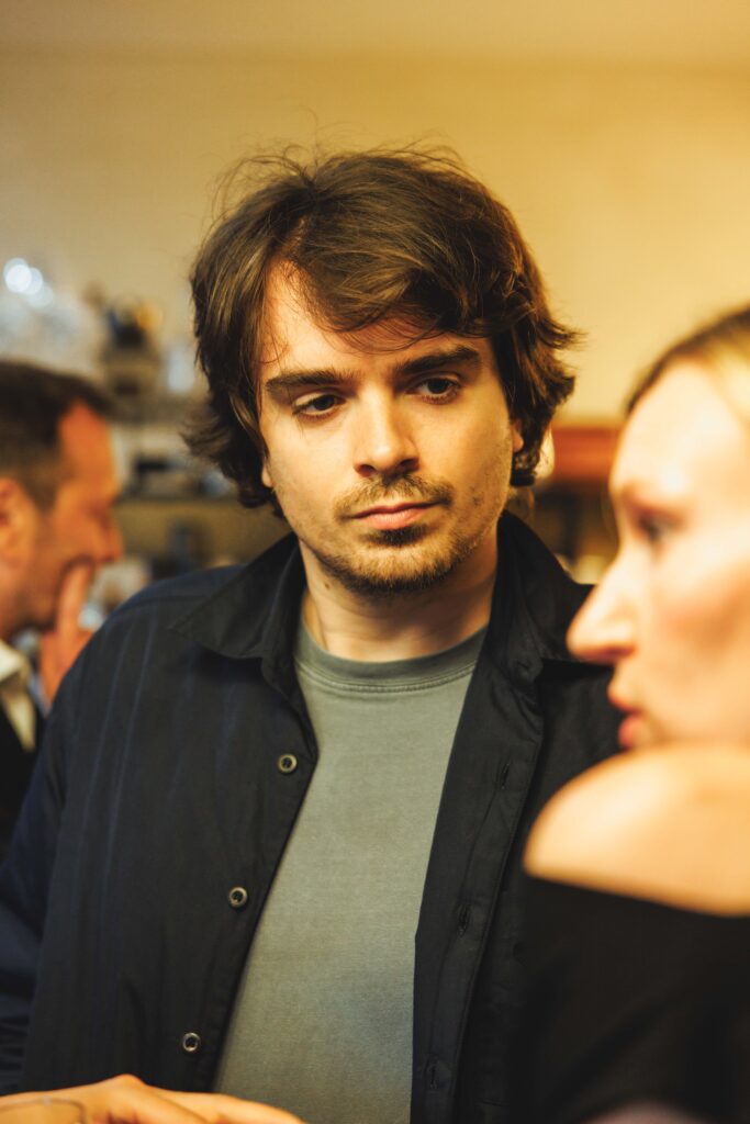 Young man with tousled brown hair gazing downward in a casual indoor gathering, wearing a gray shirt and dark jacket.