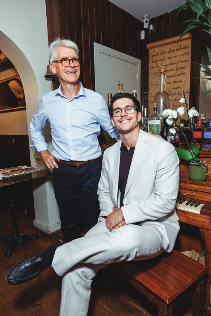 Two men posing in a cozy bar area: an older man in a blue shirt and glasses stands beside a smiling younger man in a light suit seated at a piano/bar setup with bottles in the background.