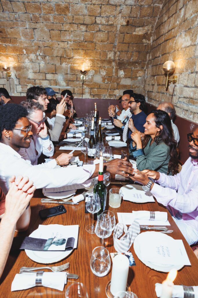 Group of diverse people seated along a long wooden table in a brick-walled, candlelit restaurant, enjoying a meal together.