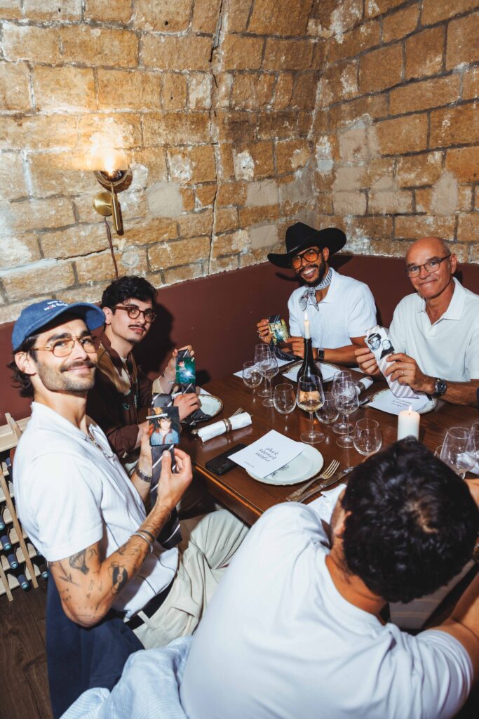 Group of men dining at a rustic brick-walled restaurant table, holding photos and smiling at the camera.