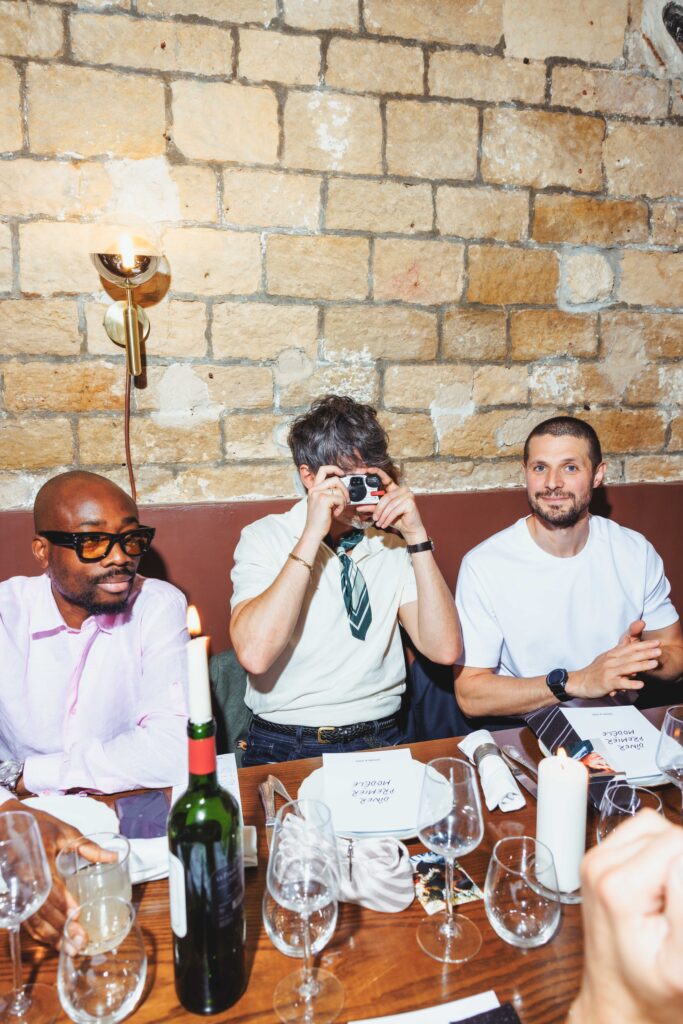Three men sit at a dining table with wine, candles, and a brick wall behind them at a restaurant party setting.