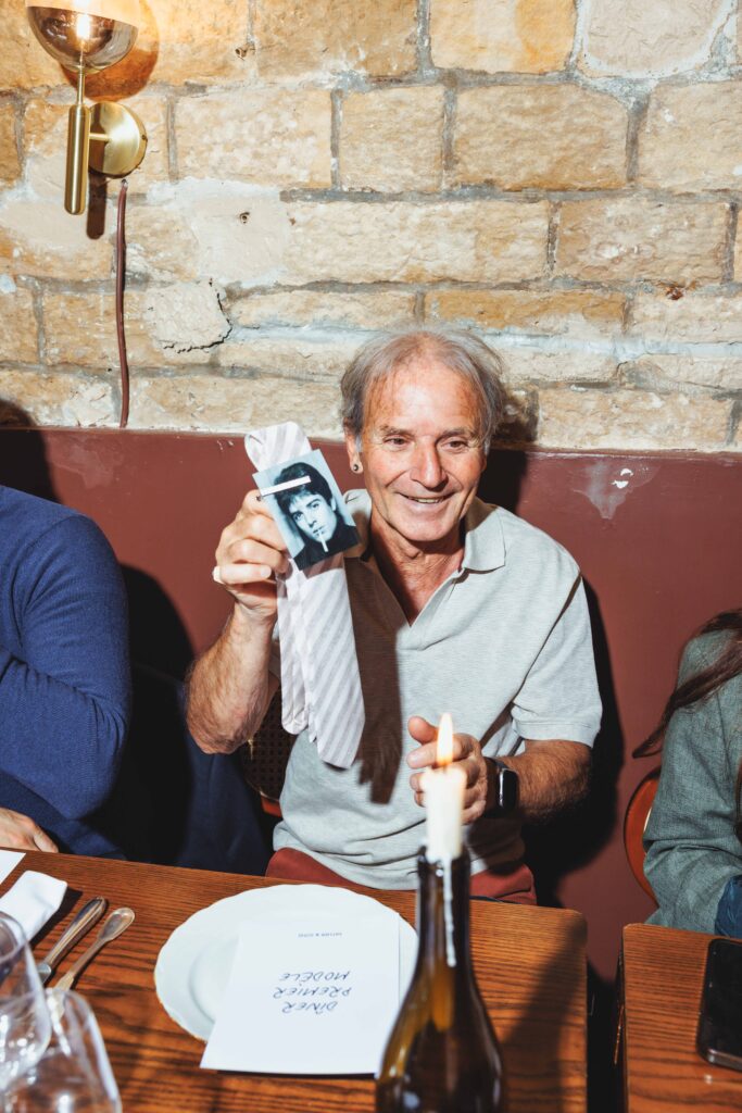 Older man smiling at a restaurant table, holding a black-and-white photo of a young man against a brick wall backdrop with a lit candle nearby