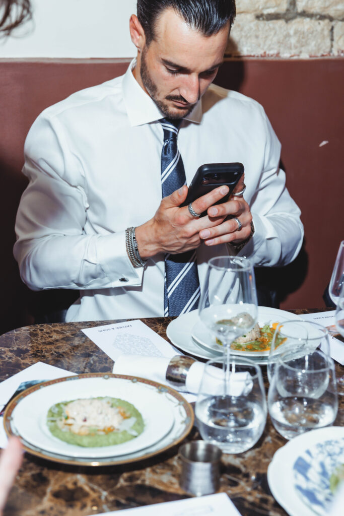 Man in a white dress shirt and striped tie sits at a restaurant table, looking at his smartphone amid plates, glassware, and menus on the table.