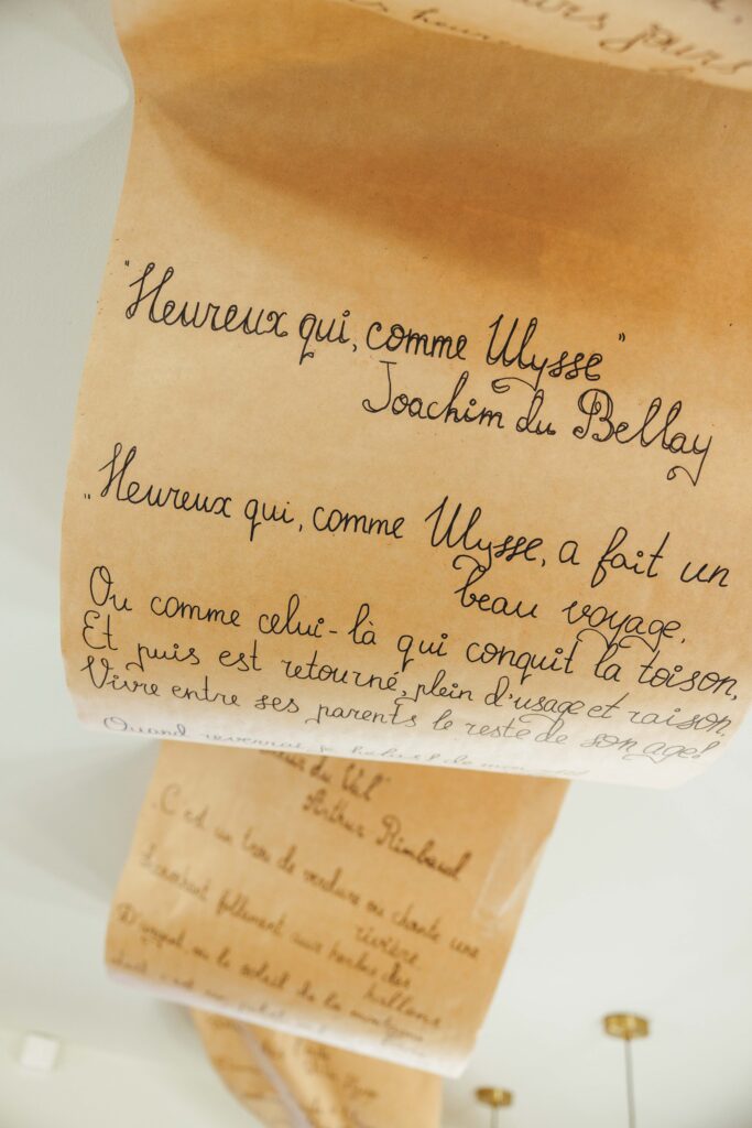 Close-up of beige parchment scrolls with cursive French handwriting, including the line "Heureux qui, comme Ulysse".