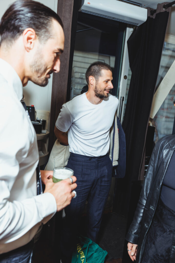 Two men in a backstage dressing area, one in a white T‑shirt smiling as the other holds a drink with foam on top nearby.