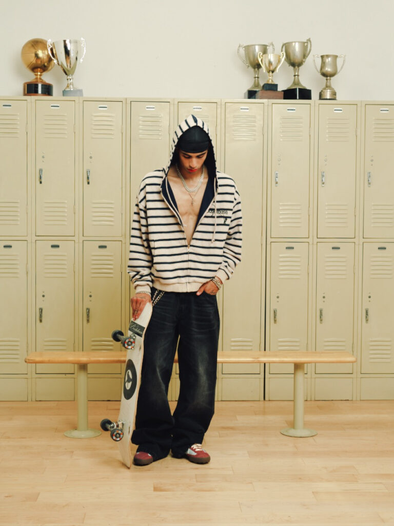 Person in a striped zip-up hoodie and dark jeans stands in a locker room, holding a skateboard, with trophies on top of beige lockers in the background.